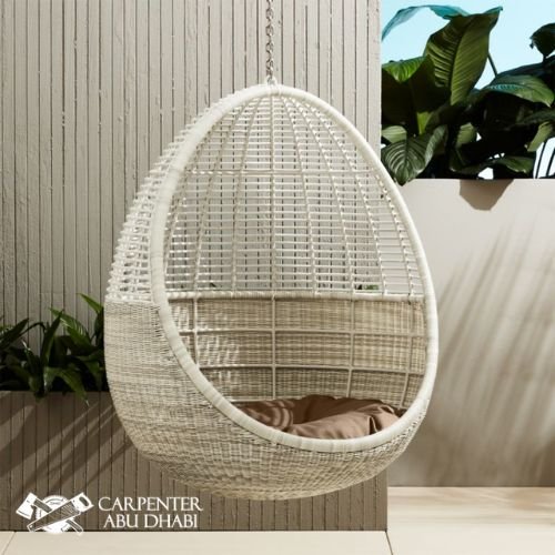 White wicker hanging egg chair with a beige cushion, suspended outdoors near plants.